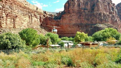 A helicopter hovers near a canyon, above green trees and houses in a valley surrounded by towering rock formations.