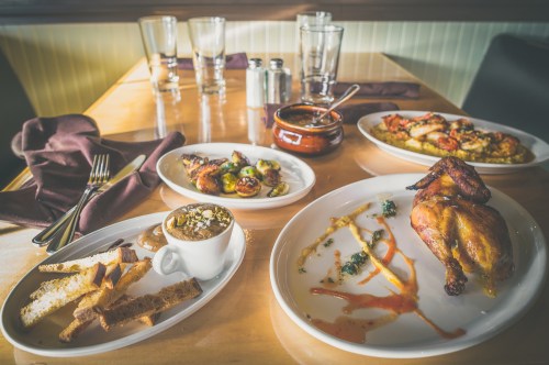 A table set with a roasted chicken, sides of vegetables, bread, and a dessert, with glasses and utensils nearby.