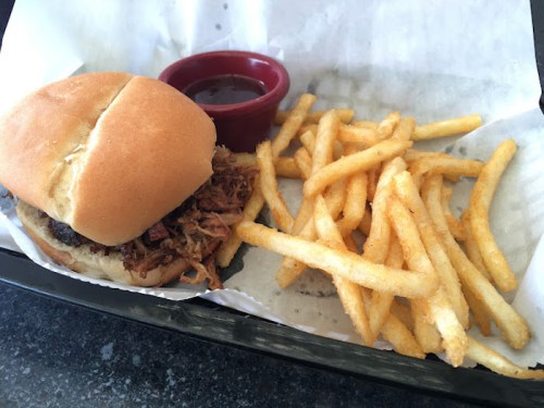 A pulled pork sandwich with a side of crispy fries and a small cup of barbecue sauce on a tray.