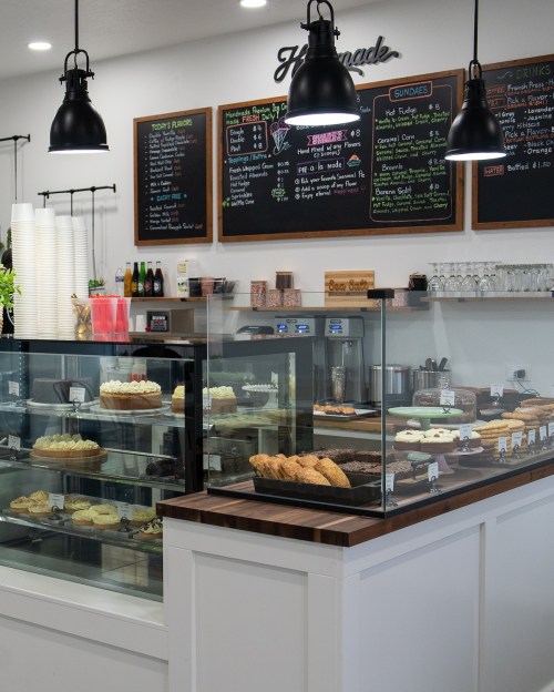 A cozy café display with glass cases showcasing desserts, a chalkboard menu, and pendant lights overhead.