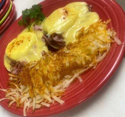 A red plate with two eggs Benedict topped with hollandaise sauce and a side of crispy hash browns, garnished with parsley.
