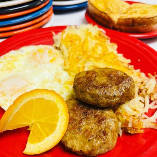 A red plate with two sausage patties, hash browns, fried eggs, and a slice of orange, with toast in the background.