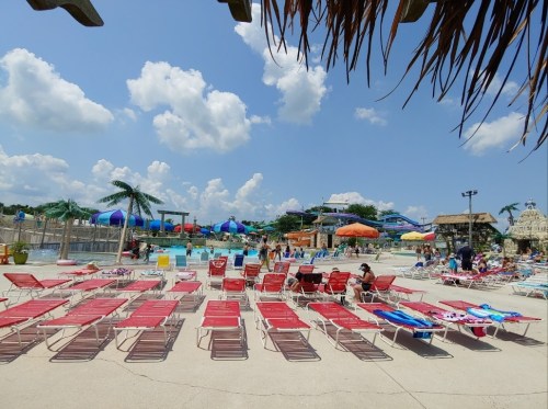 A sunny pool area with red lounge chairs, umbrellas, and water slides in the background under a blue sky with clouds.