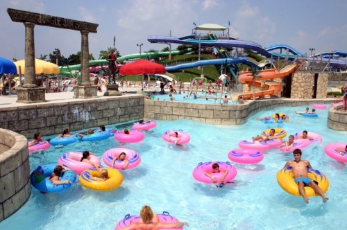 A busy water park scene with colorful inner tubes floating in a pool, surrounded by water slides and umbrellas.