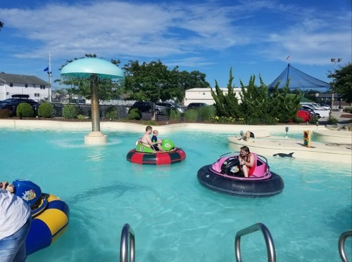 Children enjoy bumper boats in a sunny pool, surrounded by greenery and a blue sky with fluffy clouds.