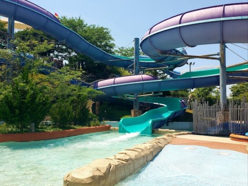 Colorful water slides twist and turn above a lazy river at a water park, surrounded by greenery and blue skies.