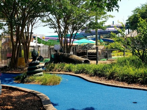 A colorful mini-golf course featuring a snake sculpture, surrounded by greenery and water slides in the background.