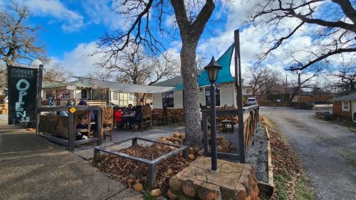 A cozy restaurant with outdoor seating, surrounded by trees, under a blue sky with scattered clouds. Sign reads "Open."