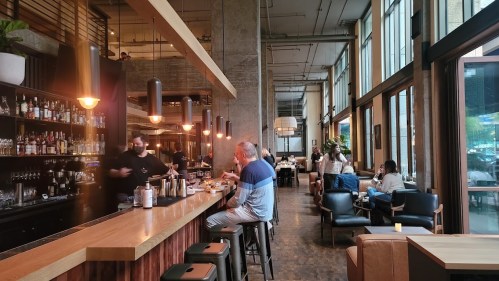 A modern bar with patrons enjoying drinks and food, featuring large windows and a stylish interior.