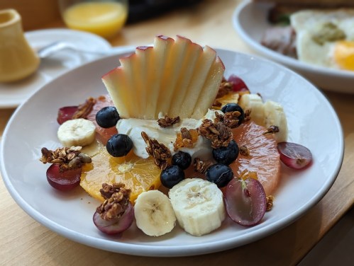A colorful fruit salad with apple slices, bananas, grapes, blueberries, and granola on a white plate.