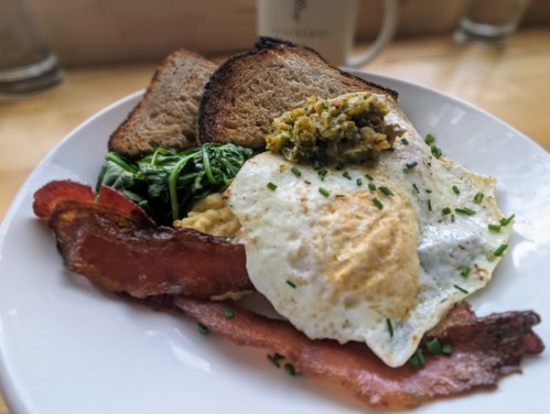A plate of breakfast featuring two fried eggs, bacon, sautéed greens, toast, and a small serving of green relish.