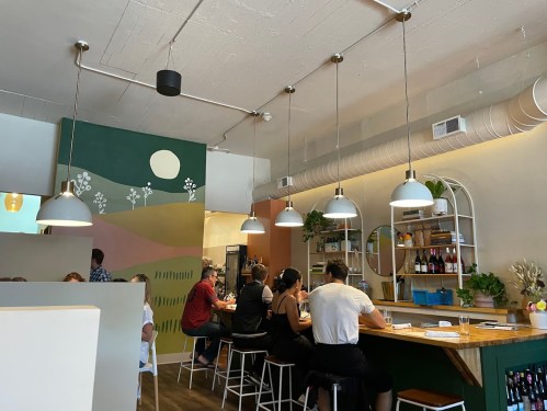 A cozy café interior with a bar, patrons seated, and a colorful mural on the wall. Pendant lights hang above the bar.