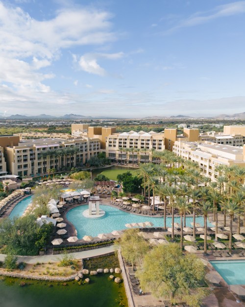 Aerial view of a resort with pools, palm trees, and mountains in the background under a blue sky.
