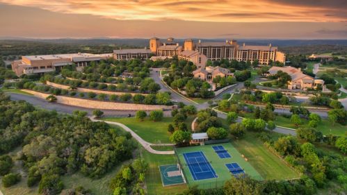 Aerial view of a resort with tennis courts, lush greenery, and a sunset sky in the background.