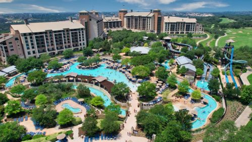 Aerial view of a resort with pools, lush greenery, and buildings in the background under a clear blue sky.