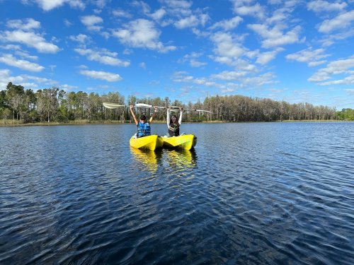 Two people in yellow kayaks raise their paddles above their heads on a calm lake under a blue sky with fluffy clouds.