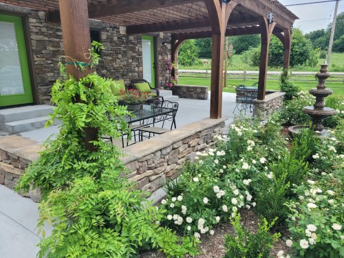 A stone patio with a wooden pergola, surrounded by lush greenery and flowers, featuring a table and chairs.