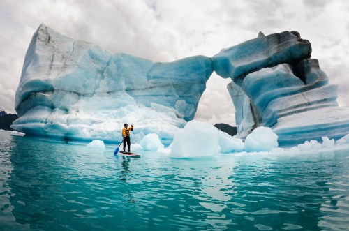A person stands on a paddleboard near a large, blue iceberg with a natural arch, surrounded by calm water.