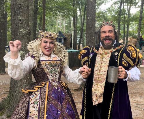A man and woman in elaborate medieval costumes pose joyfully in a forest setting, holding hands and smiling.