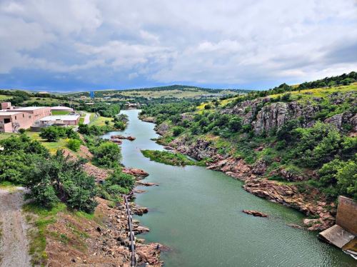 A scenic view of a winding river surrounded by rocky hills and greenery under a cloudy sky.