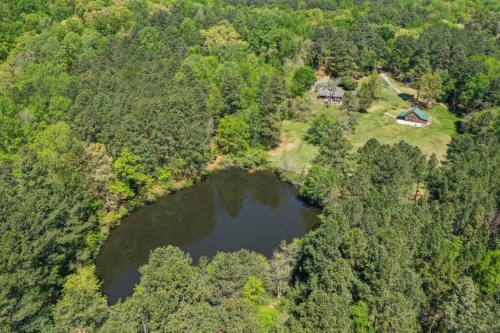 Aerial view of a serene pond surrounded by lush green trees and a small cabin in a wooded area.