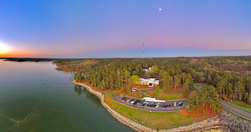 Aerial view of a lakeside area with a building, parking lot, and surrounding trees at sunset.