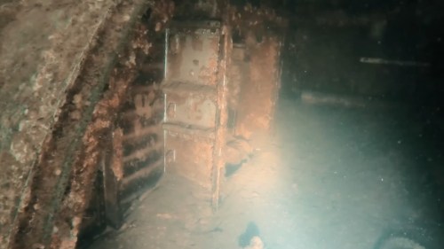 Underwater view of a rusted metal structure, possibly a door or cabinet, covered in marine growth and sediment.