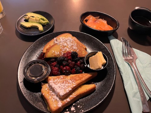 A plate of French toast topped with berries and butter, served with avocado, smoked salmon, and a small cup of syrup.