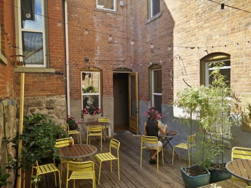 A cozy courtyard with yellow chairs, wooden tables, and potted plants, surrounded by brick walls and string lights.