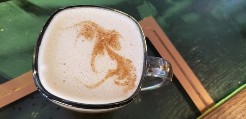 A close-up of a latte with a decorative foam design on top, served in a clear glass mug.