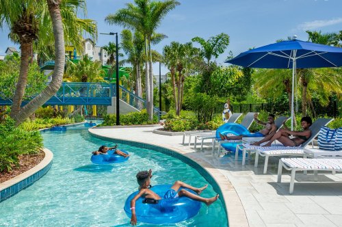 A tropical pool area with people relaxing on blue inner tubes and lounge chairs surrounded by palm trees and greenery.