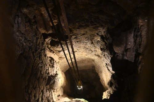 A view down a dark, rocky mine shaft with ropes hanging and a faint light at the bottom.