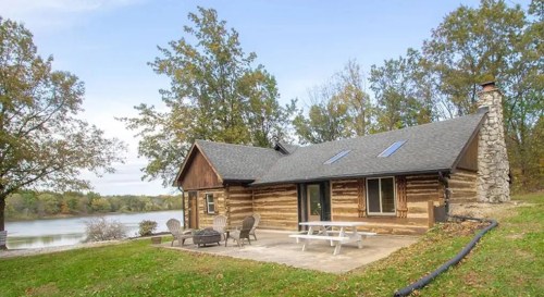 A cozy log cabin by a lake, surrounded by trees, with a stone chimney and outdoor seating area.