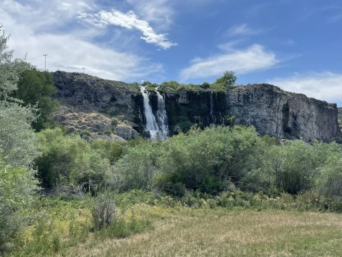 A waterfall cascades down a rocky cliff, surrounded by lush greenery and a clear blue sky.
