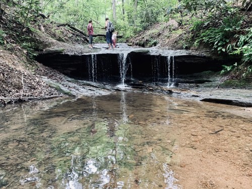 Two people stand near a small waterfall in a lush, green forest, with clear water flowing over rocks.