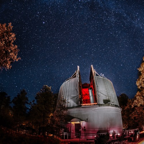 A large observatory dome under a starry night sky, surrounded by trees and illuminated by red lights.