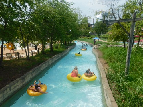 A lazy river at a water park with people floating on yellow tubes surrounded by trees and a pool area in the background.
