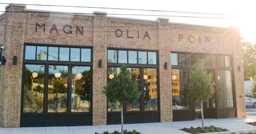 Exterior of a brick building with large windows, featuring the words "Magnolia Point" above and small trees in front.