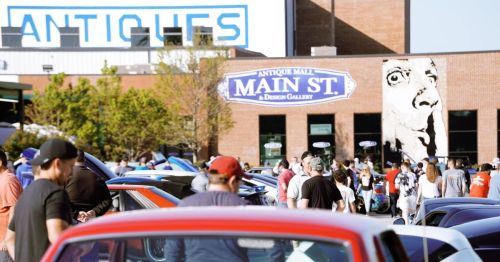 Crowd gathered outside an antique mall with vintage cars parked in front and a mural on the building.