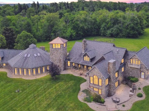 Aerial view of a large stone house with multiple sections, surrounded by lush green trees and a manicured lawn.