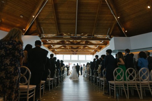 A wedding ceremony with guests seated on either side, facing a couple at the altar under a wooden ceiling.