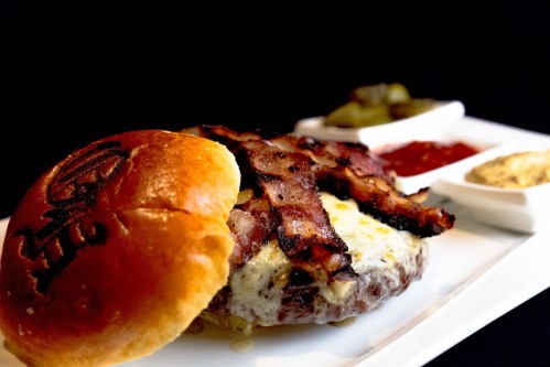 A close-up of a juicy burger topped with crispy bacon, served with condiments and pickles on a white plate.