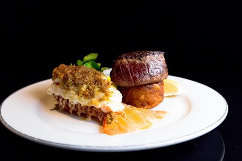 A gourmet plate featuring lobster tail, filet mignon, and a side of fried potato, garnished with lemon and greens.