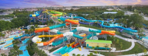 Aerial view of a colorful water park with multiple slides and pools surrounded by greenery and a sunset sky.