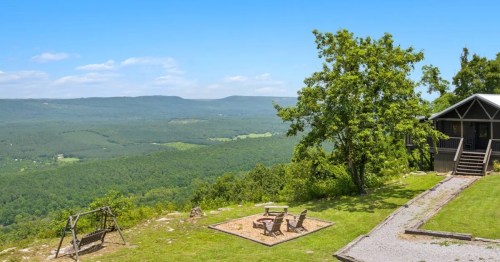 Scenic view from a hillside with a swing, fire pit, and a cabin overlooking lush green valleys and blue skies.
