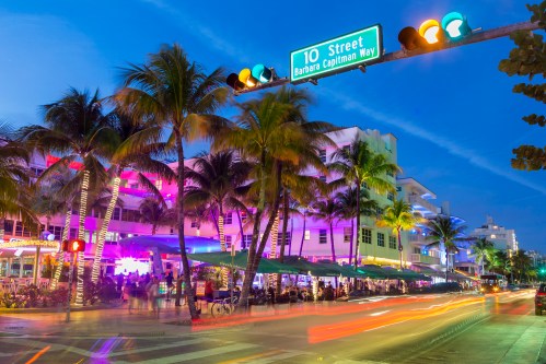 Vibrant street scene at night in Miami Beach, with palm trees, colorful lights, and a traffic signal at 10th Street.
