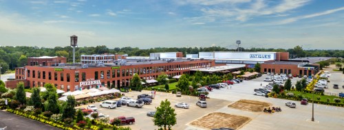 Aerial view of a brick building complex with shops, a pizzeria, and a parking lot surrounded by greenery.
