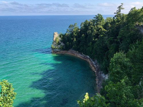 A serene coastal view featuring turquoise waters, lush green trees, and a rocky shoreline under a clear blue sky.
