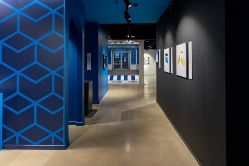 A modern gallery hallway with blue geometric wall patterns and framed artworks on a dark wall.