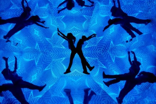 Silhouettes of dancers against a vibrant blue background with geometric patterns, creating a dynamic visual effect.